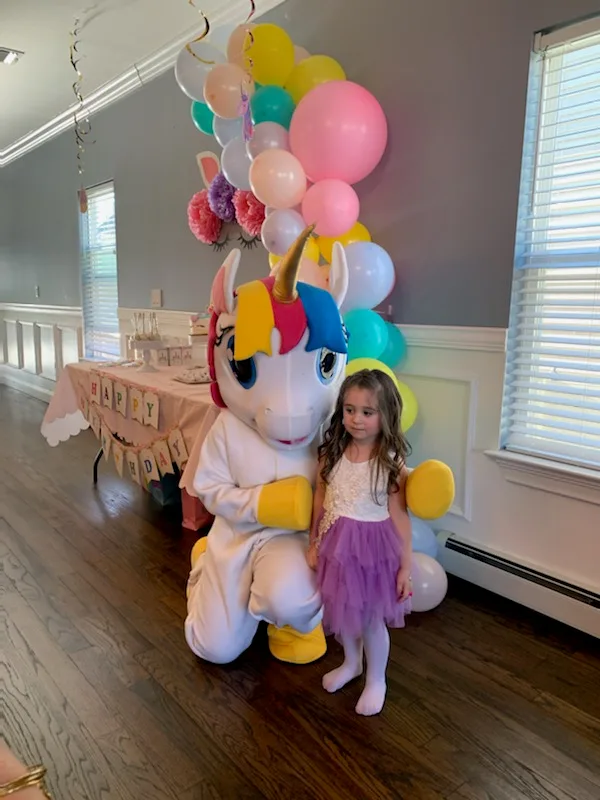A child posing with a rainbow pony mascot beside balloons