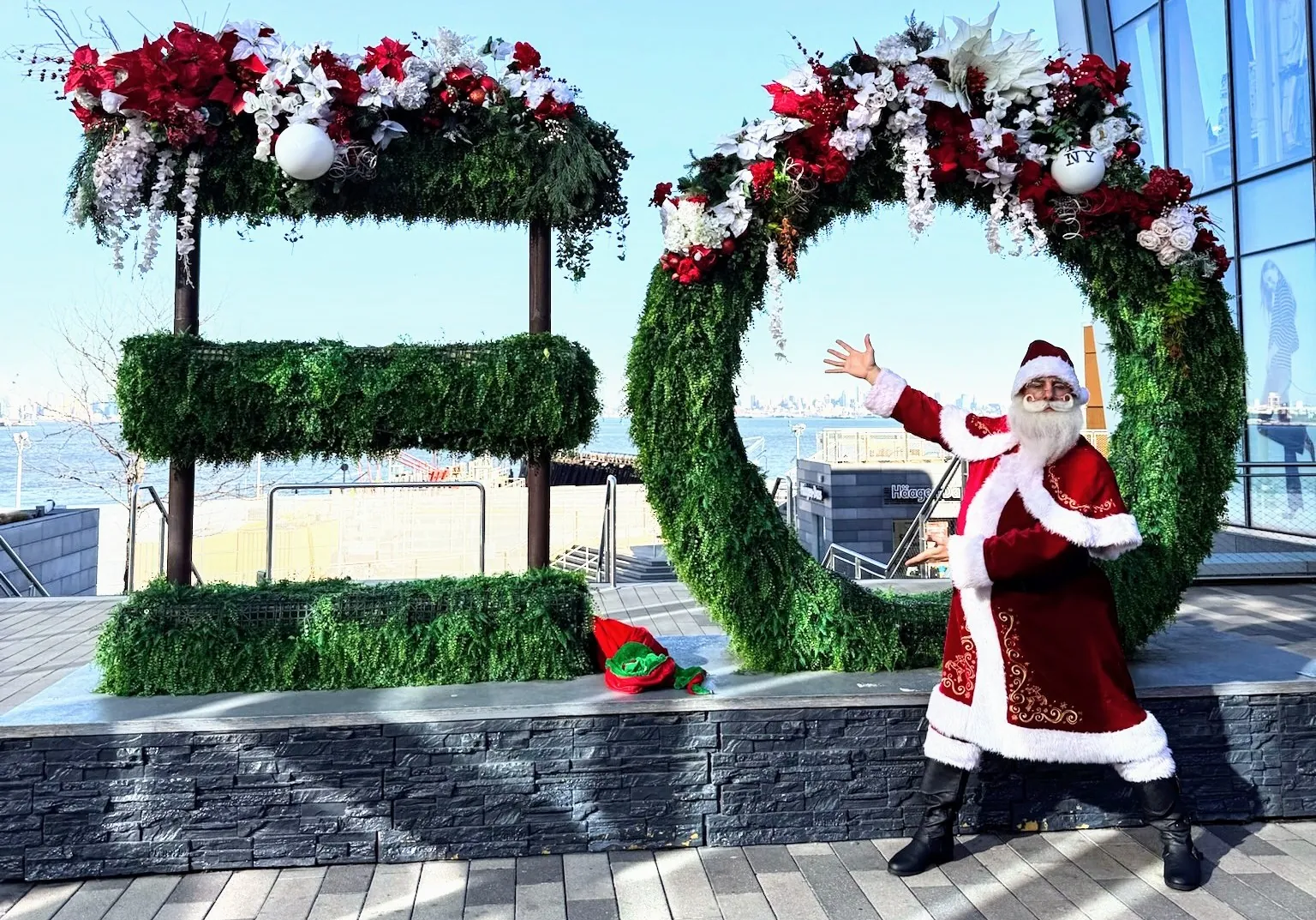 Santa waving beside oversized Christmas greenery at an outdoor holiday event